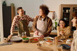 © AnnaStills - Smiling young adult Caucasian man with short hair joking and patting curly haired biracial friend on shoulder while last cutting tomatoes, young adult woman leaning on table and laughing out loud