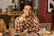 © AnnaStills - Medium close up portrait of young adult biracial man dressed in sweater with ornaments leaning his head on hand and smiling while looking at camera
