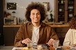 © AnnaStills - Medium close up portrait of curly haired young adult biracial man holding cutlery looking at camera and smiling