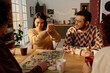© AnnaStills - Group of friends looking at young adult woman who smiling while shaking dice, they playing board game at home