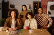 © AnnaStills - High angle portrait of biracial company of friends sitting at table with board game and aroma candles, looking at camera while posing for group photo