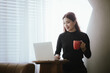 © Witoon - A young woman is relaxing in a cozy coffee shop, smiling as she sips her coffee. She uses her laptop and smartphone to multitask, whether it's reading a book or surfing the web, combining her modern l