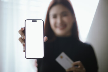 Young Asian woman smiling while using smartphone and holding credit card to buy things online. Image highlights e-commerce, finance, technology and convenience of digital transactions.