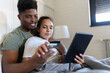 © lordn - A young couple sits together on a bed, holding a credit card and tablet, smiling as they engage in online shopping in a cozy, well-lit bedroom