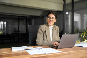 Smiling professional financial it specialist latin hispanic business lady working on laptop pc sitting in office. Middle eastern indian woman dreaming using computer technology app for work online