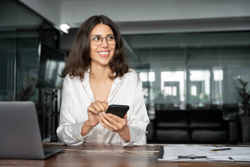 Middle aged Hispanic business manager ceo using cell phone mobile app, laptop. Smiling Latin or Indian mature woman businesswoman holding smartphone sit in office working online on gadget, copy space.