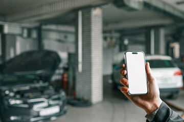 Fototapete - Hand holding a smartphone with a blank white screen in a car repair workshop, featuring blurred vehicles and garage equipment in the background