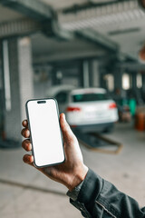Fototapete - Hand holding a smartphone with a blank white screen in a car repair workshop, featuring blurred vehicles and garage equipment in the background