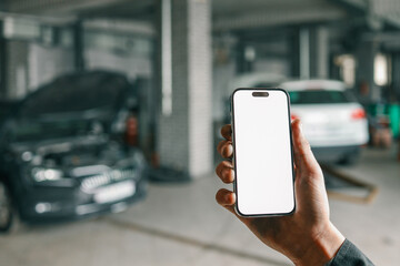 Fototapete - Hand holding a smartphone with a blank white screen in a car repair workshop, featuring blurred vehicles and garage equipment in the background