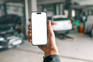 Fototapete - Hand holding a smartphone with a blank white screen in a car repair workshop, featuring blurred vehicles and garage equipment in the background
