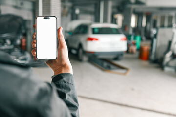 Fototapete - Hand holding a smartphone with a blank white screen in a car repair workshop, featuring blurred vehicles and garage equipment in the background