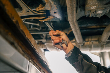 Aufkleber - Mechanic's hand holding a wrench, working underneath a car on a lift. The view shows a close-up of the suspension and exhaust system in a garage setting