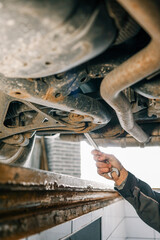 Aufkleber - Mechanic's hand holding a wrench, working underneath a car on a lift. The view shows a close-up of the suspension and exhaust system in a garage setting