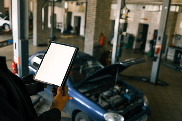Fototapete - Mechanic holding a tablet with a blank screen in a car repair shop. A blue car with an open hood is in the background, and the workshop has visible car lifts and tools