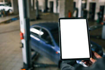 Fototapete - Mechanic holding a tablet with a blank screen in a car repair shop. A blue car with an open hood is in the background, and the workshop has visible car lifts and tools