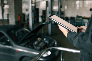 Fototapete - Mechanic holding a tablet in an automotive workshop, inspecting car diagnostics and maintenance information, with a blurred repair area in the background
