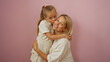© Krakenimages.com - Blonde woman embracing her daughter who is giving her a kiss on the cheek in front of a pink background, conveying love and family bonding.