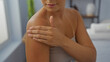 © Krakenimages.com - Young woman with hispanic ethnicity in a spa room applying lotion to her shoulder, showcasing a wellness and beauty routine indoors.