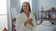 © Krakenimages.com - Woman relaxing in a spa room, holding a lotion bottle, wearing a bathrobe, smiling and enjoying the serene indoor spa setting with shelves and a massage bed in the background