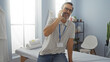 © Krakenimages.com - Man talking on phone while sitting in a bright clinic room with a massage table, wearing glasses and a lanyard, surrounded by indoor plants and modern decor.