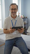 © Krakenimages.com - Handsome middle-aged hispanic man wearing glasses and a lanyard, holding a tablet in a modern clinic interior, seated casually, exuding professionalism and warmth.