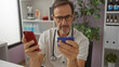 © Krakenimages.com - Mature hispanic man in a clinic room analyzing a credit card with a smartphone, surrounded by medical equipment and plants, suggesting a workplace setting.