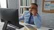 © Krakenimages.com - Young man with braids working in an office wearing a blue shirt and glasses, sitting at a desk with computer and notebooks, appearing thoughtful and focused.