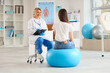 © Pixel-Shot - Young woman on fit ball visiting mature female osteopath with clipboard in clinic