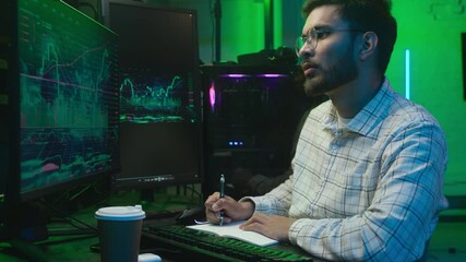 Indian Arabian man writing notes notebook computer pc monitor freelancer crypto trading chart screen male programmer guy IT broker analyst write bitcoin online currency working results neon e-commerce