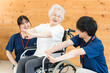 © buritora - リハビリで体操をする車椅子に乗ったシニア女性と理学療法士・作業療法士の男女スタッフ