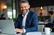 © SimpleDesignStudio - Professional Mature Man Working on Laptop in Modern Office Environment, Smiling and Engaged in Work with Colleagues in Background