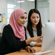 © Paintbrush - Two women collaborating on a computer, one wearing a pink hijab, in a modern office setting.