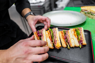 Wall Mural - A chef arranging freshly made sandwiches on a serving tray. The sandwiches feature layers of meats and vegetables, showcasing a colorful and appetizing presentation in a professional kitchen.