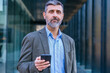 © alfons - Confident businessman using smartphone outdoors looking at camera,
