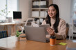 © Tj - Smiling young Asian businesswoman working on a laptop while reading a book in a modern office, enjoying takeaway coffee surrounded by sticky notes on her desk