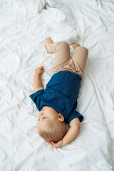 Wall Mural - A blond one-year-old boy in a blue T-shirt lies on a bed with white sheets. The baby plays while sitting in bed. A blue-eyed little boy.