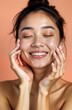 © miss irine - Young Asian woman with healthy skin. Portrait of beautiful girl with natural makeup. Touches face gently. Happy expression, glowing skin. Studio shot against peach background. Skincare, cosmetics