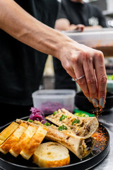 Wall Mural - A hand sprinkles seasoning over a gourmet dish featuring roasted bone marrow served with slices of toasted bread and garnishes, capturing a moment of culinary expertise in a kitchen setting.