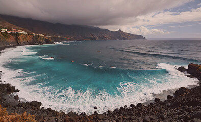 Poster - The ocean is calm and blue, with a few small waves breaking on the shore. The sky is cloudy, giving the scene a moody and serene atmosphere