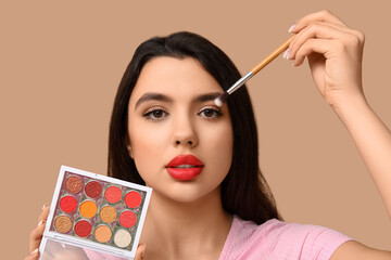 Wall Mural - Beautiful young woman with palette of eyeshadows and makeup brush on brown background