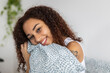 © Xavier Lorenzo - Young latin american woman smiling and hugging a pillow in bed, enjoying a relaxing morning at home