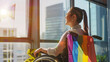 © DEIB Visuals - Happy gay asian woman in a wheelchair with a rainbow flag looking out of office window. Smiling lesbian businesswoman celebrating pride month 2025