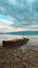 Naklejka na meble Fishing boat on the edge of the lake surrounded by snowy mountains on a cloudy sunset. The sky and the lake are painted blue, the sun makes you feel its warmth with its reflections on the lake. 