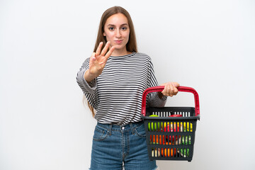 Young Rumanian woman holding a shopping basket full of food isolated on white background happy and counting three with fingers