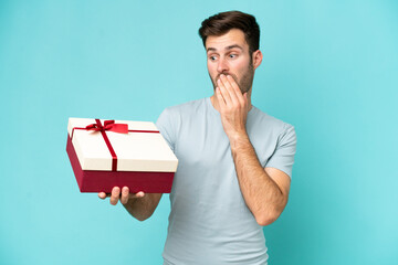 Wall Mural - Young caucasian man holding a gift isolated on blue background with surprise and shocked facial expression