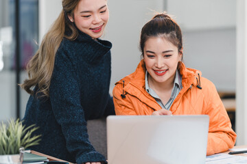 Wall Mural - Two women collaborating at desk, smiling while looking at laptop screen, creating positive and productive atmosphere in modern office setting