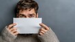© Роман Варнава - Young man holding blank paper in front of his face against a gray background in a casual setting