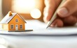 © apichat - A miniature orange house sits on paperwork as a hand holds a pen, symbolizing real estate transactions or home buying processes.
