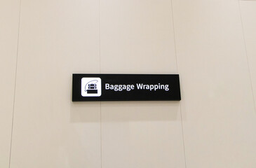 Black signs with white letters and Luggage symbol for Baggage wrapping service at International Airport. On white wall is background with light and shadow from light. Inform tourists about places.