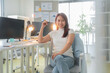 © whyframeshot - Asian young woman sitting in modern office, smiling confidently while working at her desk. workspace features computers, plants, and bright lighting, creating productive atmosphere